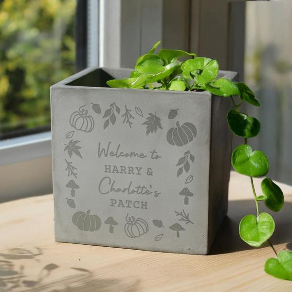 Grey square concrete plant pot engraved with pumpkins and the words Welcome to Harry & Charlotte’s Patch, sitting on a wooden table holding a trailing green plant.