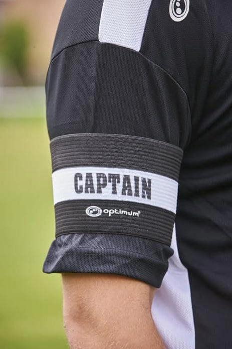 Close-up of black Optimum CAPTAIN armband worn on a player's arm over black kit on grass - Optimum