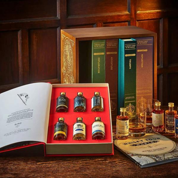 A luxurious, open wooden bookcase revealing five linen-bound books titled 'Exploration', 'Adventure', 'Triumph', and 'Reflection'. The front book is open, showcasing six small premium whisky bottles on a red felt tray. Beside the bookcase sits a whisky glass with amber liquid, an additional small bottle, and a circular 'Whisky Tour of the World' guidebook, all arranged on a dark wooden table.