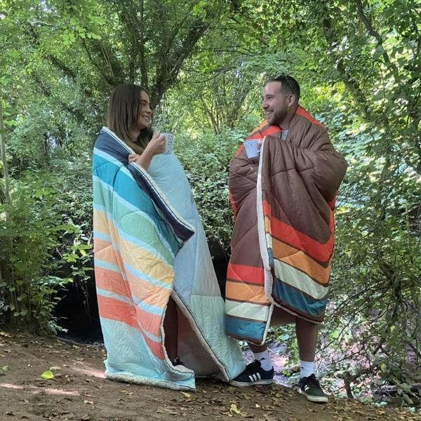 Tonal treetops and vivid spruce Insulated Outdoor Blanko been worn by a young couple enjoying the countryside with the outdoor blanket, wrapped around them.