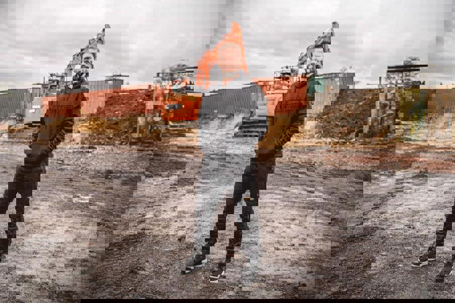 WorkTough Mens Cargo Stretch Fit Cordura Work Trouser Fleece Jogger, man standing on a construction site in front of heavy machinery wearing black work trousers and hoodie.