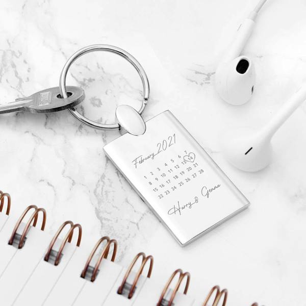 A rectangular silver metal keyring lying on a white marble surface, displaying a calendar page for "February 2021" with the 14th circled and the names "Harry & Genna" engraved below. The keyring is surrounded by a spiral notebook and wired earbuds.
