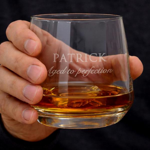A hand holding a heavy-based glass tumbler filled with whisky. The engraving features the name "PATRICK" in block capitals with the phrase "Aged to perfection" in an elegant script beneath it.
