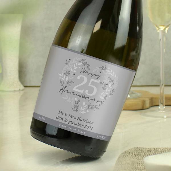 A tilted bottle of personalised 25th Silver Wedding Anniversary Prosecco on a table next to a champagne flute, with the custom label showing an example reading Mr & Mrs Harrison 19th September 2024.