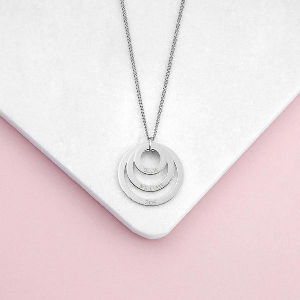 A close-up shot of a three-tier silver nesting family ring pendant necklace engraved with the names 'OLLIE', 'WILLIAM', and 'ZOE' displayed vertically down the face of each concentric loop, resting neatly on a cornered white tile over a soft pink background.