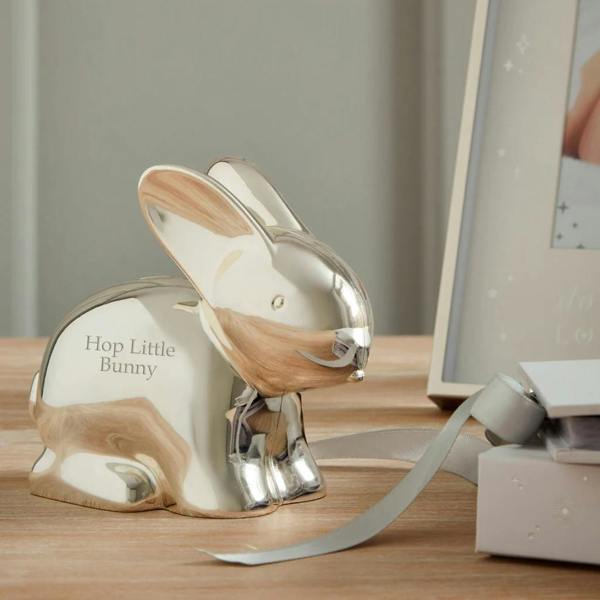 A polished silver-plated bunny money box engraved with Hop Little Bunny sitting on a wooden tabletop next to a white ribbon and a gift box.