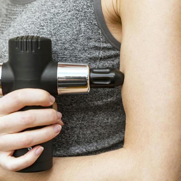 A person in a gray tank top uses the Mini Deep Tissue Massage Gun on their bicep, applying the device to their upper arm for deep tissue relief.