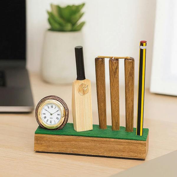A wooden cricket-themed desktop clock and pencil stand showing a miniature bat, stumps, and a small round analogue clock with Roman numerals on a green felt base, holding a yellow and black pencil on a modern desk setup.