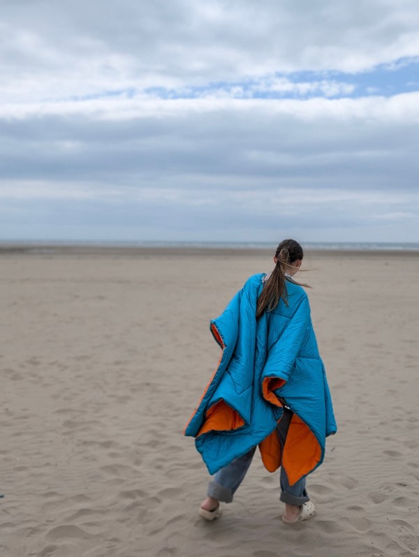 A lady walking along the beach wearing a blue/Orange reversible Blanko as a poncho.