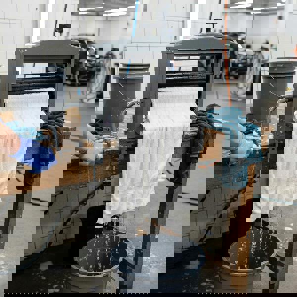 Manual hand wringer with a white towel being squeezed, draining water into a bucket on a wooden table in a clean workspace