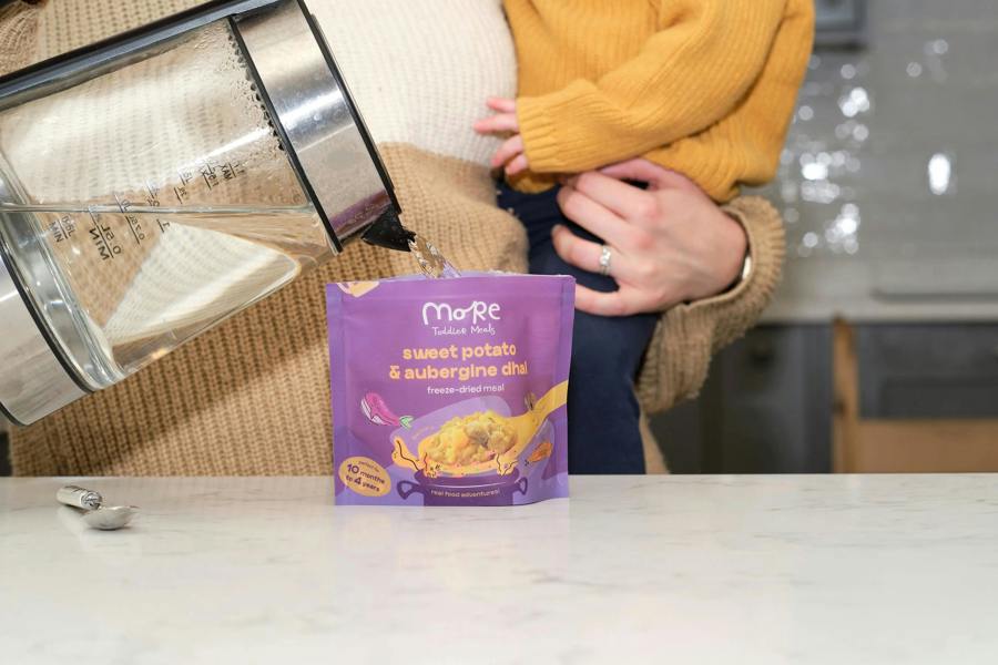 A Mum holding a toddler whilst preparing a more toddler meal with hot water. 