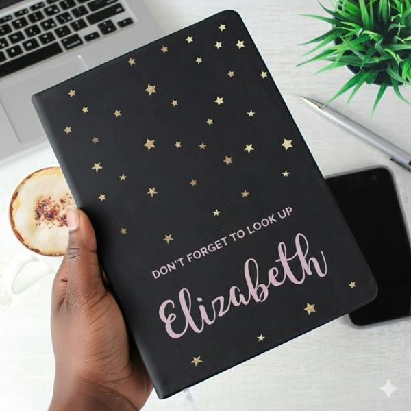 A hand holding a black A5 hardback notebook with gold foil star patterns. The cover features the text Don't forget to look up followed by the name Elizabeth in a pink script font.