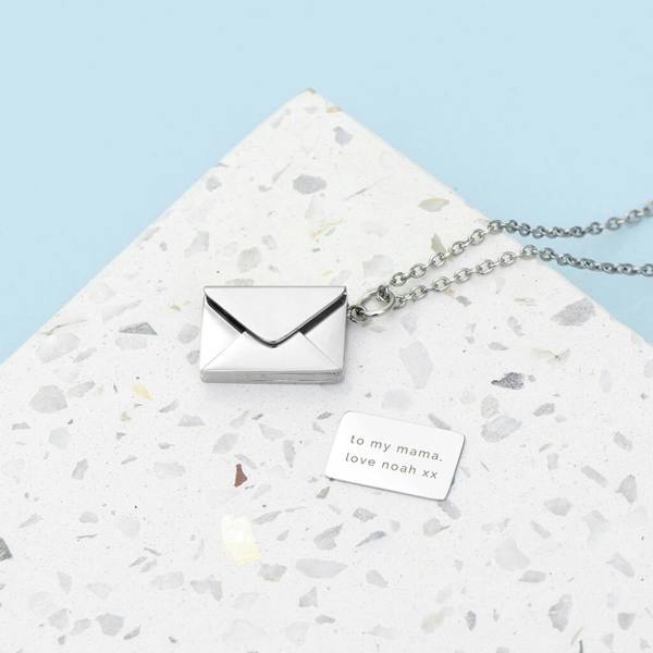 A silver envelope necklace on a speckled terrazzo surface with its small hidden card pulled out, reading 'to my mama. love noah xx'.