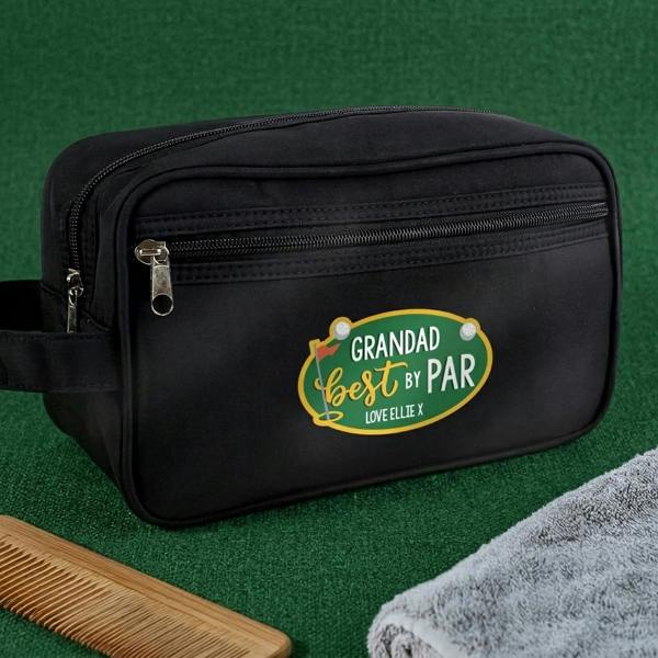 A black canvas wash bag with a custom green 'Grandad Best By Par' badge resting on a textured green surface next to a grey towel and a wooden comb.