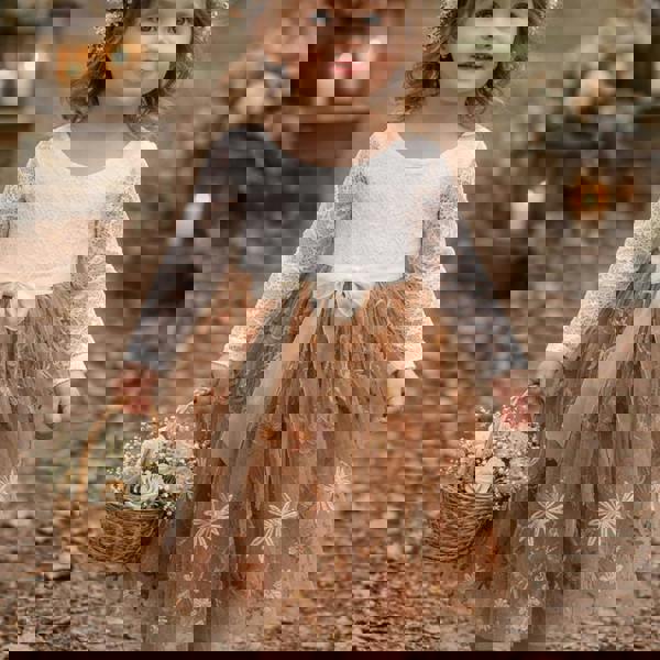 Young girl in a white top and brown floral dress holding a basket of flowers, standing outdoors with candles and flowers in the background.