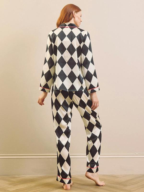 A woman stands barefoot against a beige wall, viewed from the back, wearing a matching long-sleeve pyjama set with a black and cream harlequin diamond pattern and thin pink piping along the collar, cuffs, and hem.