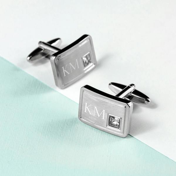 A pair of rectangular, brushed silver-tone cufflinks with a small crystal inset, engraved with the initials "KM" and displayed on a light surface.
