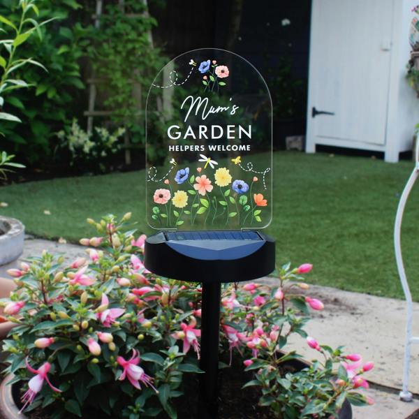 A daytime close-up of an unlit clear arched acrylic solar garden light stake. The panel features a vibrant printed wildflower meadow design with a dragonfly and bees surrounding the custom white text 'Mum's GARDEN, HELPERS WELCOME'. The black solar-cell base is staked directly into a potted pink and purple fuchsia plant, with a blurred green lawn and white garden shed in the background.