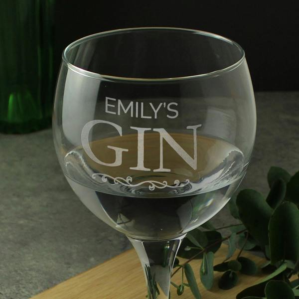 A close-up shot of a personalised stemmed gin balloon glass by Abode Home & Gifts, showing the frosted laser-engraved text "EMILY'S GIN" above an elegant scroll, filled with a clear drink on a wooden board.