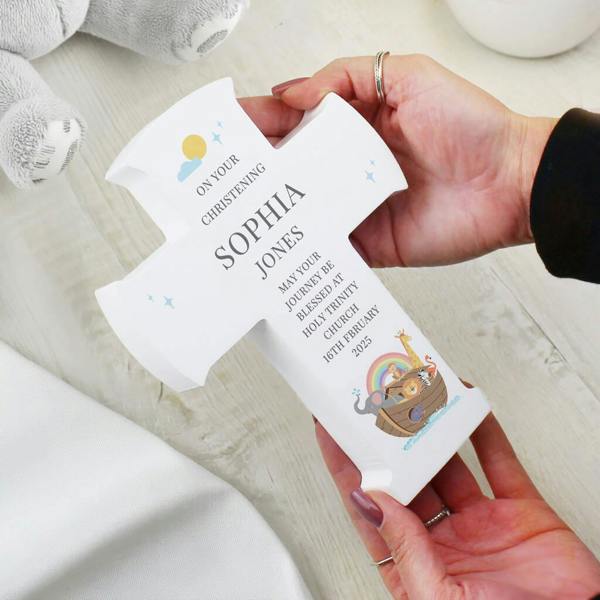 A white wooden freestanding cross reading On Your Christening Sophia Jones with a colorful Noah's Ark illustration held in two hands.