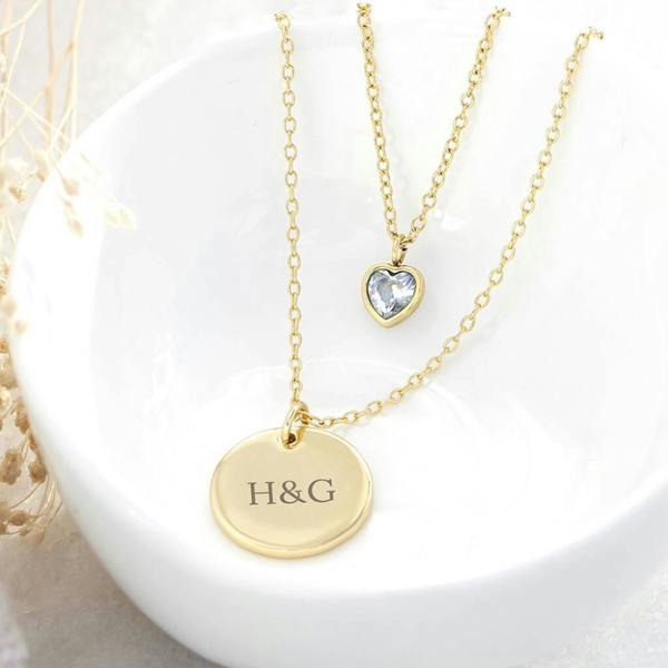 A personalised layered gold necklace featuring a small crystal heart charm and a circular gold disc engraved with the initials H&G.