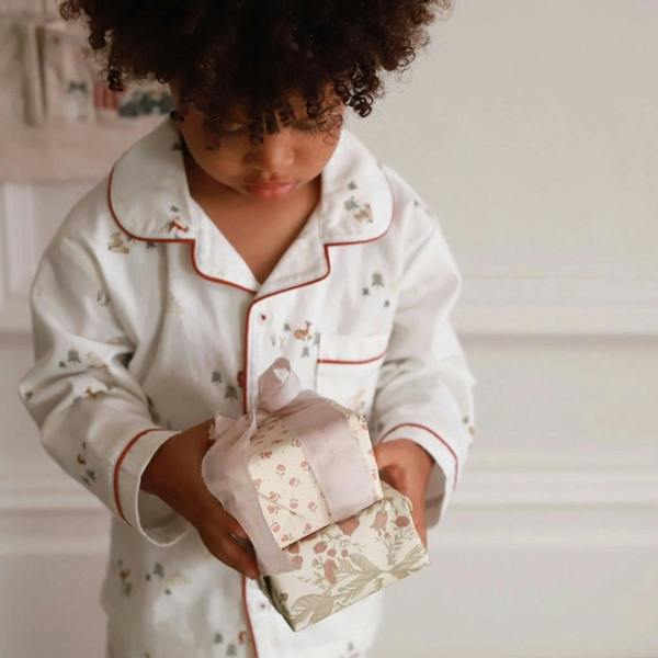 A close-up of a young child holding small wrapped presents, wearing white organic cotton festive pyjamas with red piping details.
