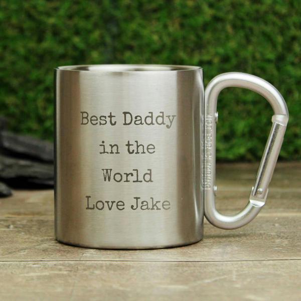 A brushed metal camping mug with an integrated silver carabiner handle, sitting on a rustic wooden surface. The front is professionally engraved with the text 'Best Daddy in the World Love Jake'.