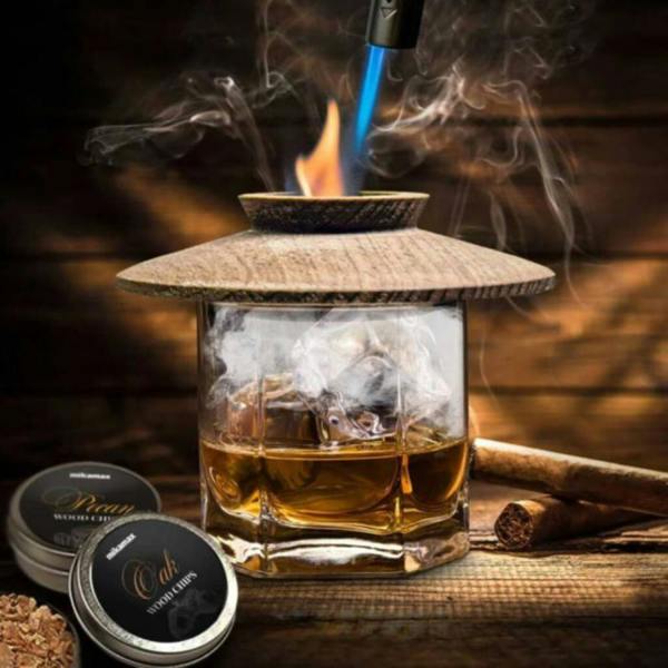 A professional whisky smoker kit in action, featuring a wooden chimney top placed over a glass of amber whisky. A blue flame from a torch ignites wood chips, creating a swirl of natural smoke inside the glass. Two tins of wood chips—Oak and Pecan—sit beside it on a rustic wooden table, with a cigar resting nearby to complete the aesthetic.