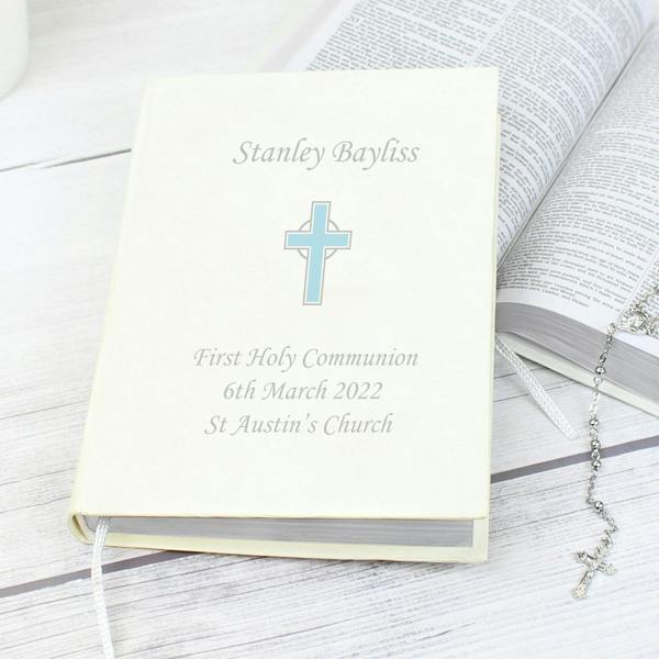 The front cover of the Personalised Eco Friendly Blue Cross Holy Bible from Abode Home & Gifts customised for Stanley Bayliss's First Holy Communion, resting at an angle on a white wood surface next to an open Bible and a silver rosary bead necklace.