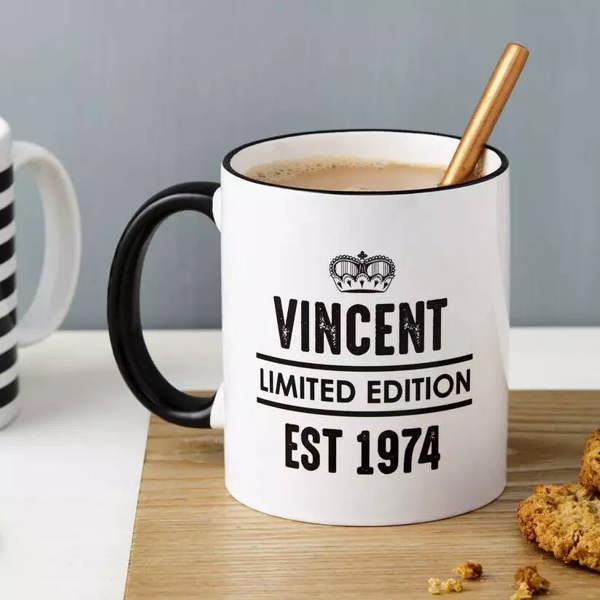 A personalised white ceramic mug with a black handle and rim, featuring the text Vincent Limited Edition Est 1974 beneath a crown graphic, styled on a wooden board next to biscuits.
