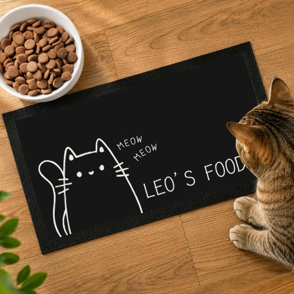 Top-down view of a black personalised cat feeding mat reading LEO’S FOOD next to a bowl of dry cat food and a tabby cat.