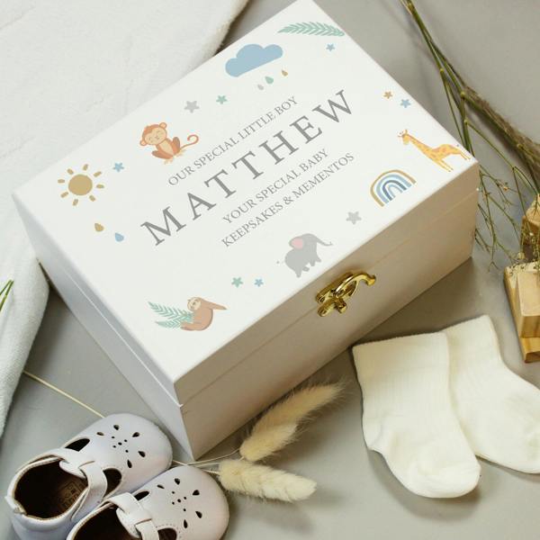 A rectangular hand-painted white wooden keepsake box with a gold metal clasp sits angled on a grey surface. The lid is printed with safari animals including an elephant, giraffe, monkey, and sloth around the capitalised custom text 'OUR SPECIAL LITTLE BOY, MATTHEW, YOUR SPECIAL BABY, KEEPSAKES & MEMENTOS'. White baby shoes and cream socks sit in the foreground.