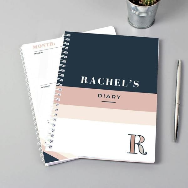 A spiral-bound Personalised A5 Diary with a navy and pink geometric cover custom-printed with Rachel's Diary and a large initial R next to a silver pen.