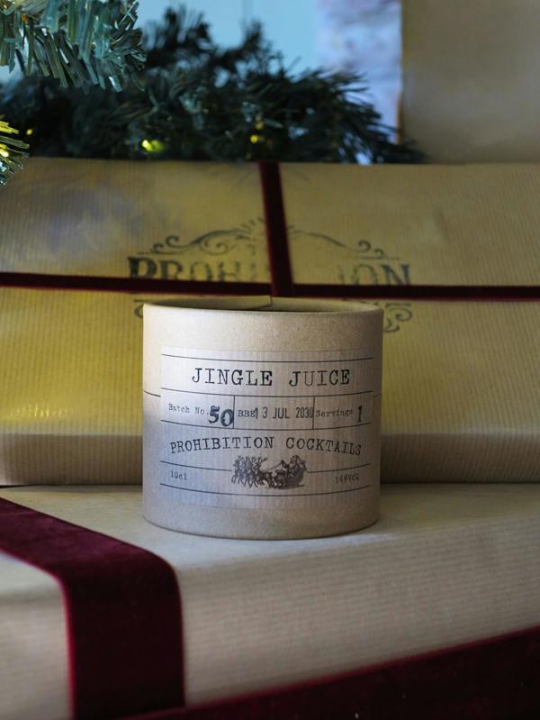 A cylindrical candle labeled 'JINGLE JUICE' and 'PROHIBITION COCKTAILS' sits on top of wrapped gift boxes with a dark red ribbon, with pine branches in the background.