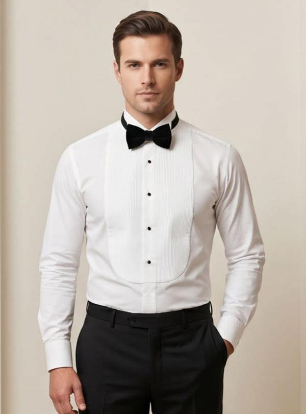 A man wearing a white tuxedo shirt with a black bow tie and black trousers, standing against a plain beige studio background.