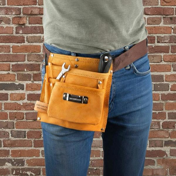 A close-up of a person wearing a tan, heavy-duty leather tool belt over blue jeans against a brick wall. The belt has six pockets filled with tools, including a wrench, and a leather tab engraved with "DAD CAN FIX ANYTHING".