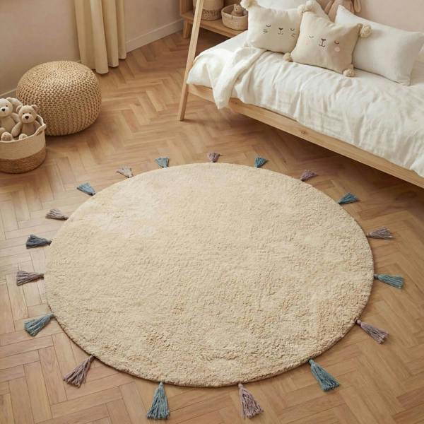 Round beige rug with tassels on a wooden floor in a bedroom setting.