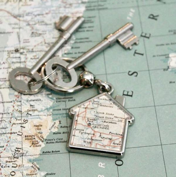 A silver house-shaped keyring displaying a section of a vintage map, lying on a large faded world map background with two skeleton keys attached to the ring.