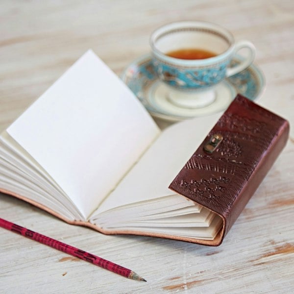 The open Indra Medium Embossed Leather Journal with its rich brown cover sits elegantly on the wooden table alongside a pencil and a teacup on a saucer.