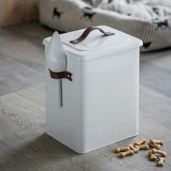 A medium-sized white chalk-coloured powder-coated steel pet food bin with a brown leather lid handle and side scoop, placed on a wooden floor next to bone treats with a pet bed in the background.
