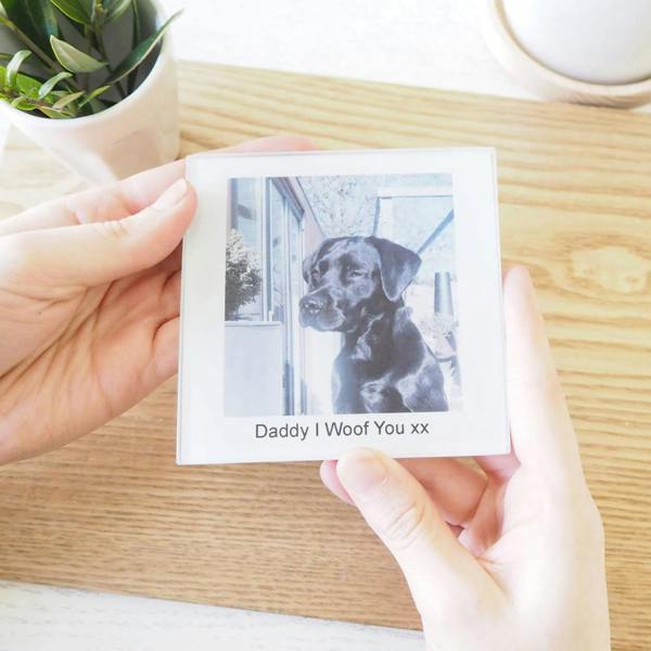 A person's hands gently hold a square acrylic personalised coaster featuring a photo of a black Labrador dog looking out a window. Below the photo, the white background is printed with the personalised text "Daddy I Woof You xx". A wooden tabletop and a green plant are visible in the background.