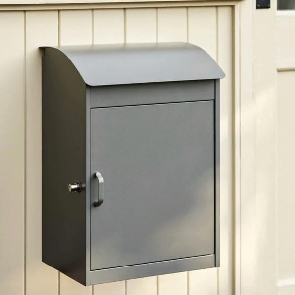 A charcoal grey metal parcel box mounted on a cream wooden wall with a secure lock and handle.