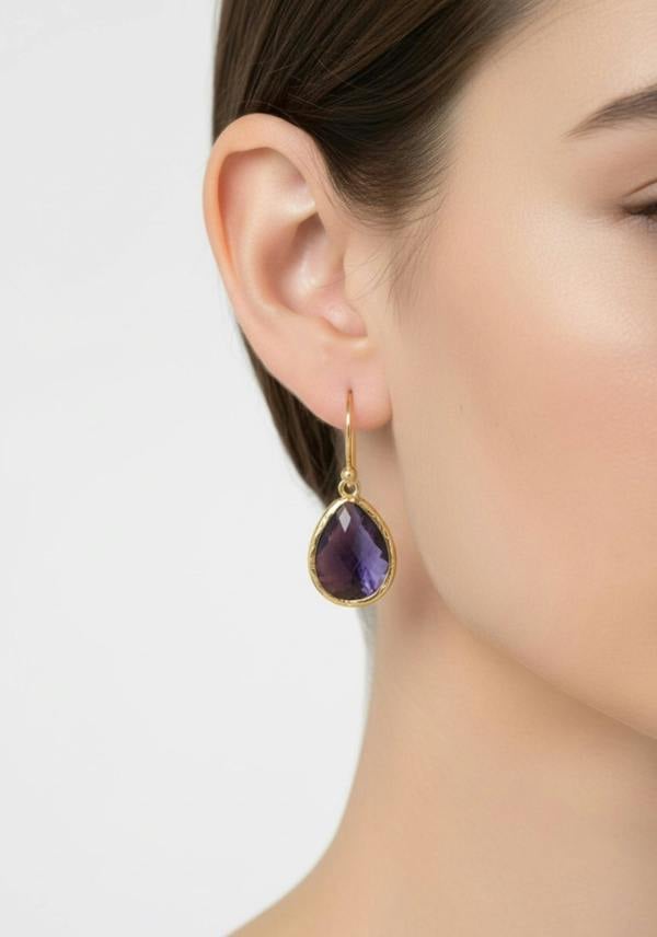Close-up of a woman wearing a gold earring with a purple gemstone on a neutral background