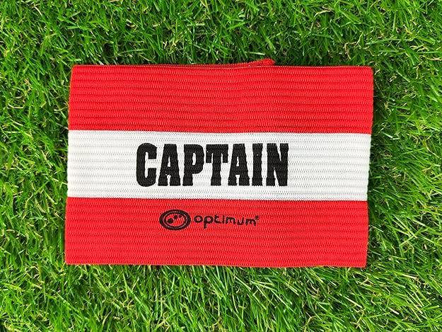 Red Optimum captain's armband with white CAPTAIN stripe and logo laid flat on grass - Optimum