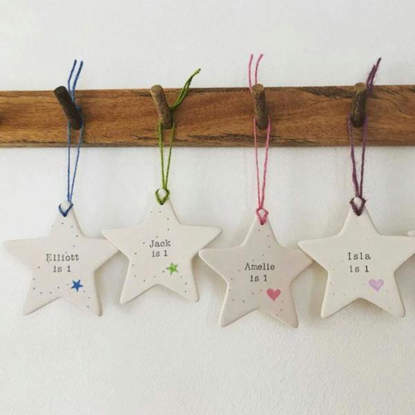 Four personalised white ceramic star ornaments hanging from a wooden rack, hand-stamped with names like Elliott, Jack, Amelie, and Isla for their 1st birthday.