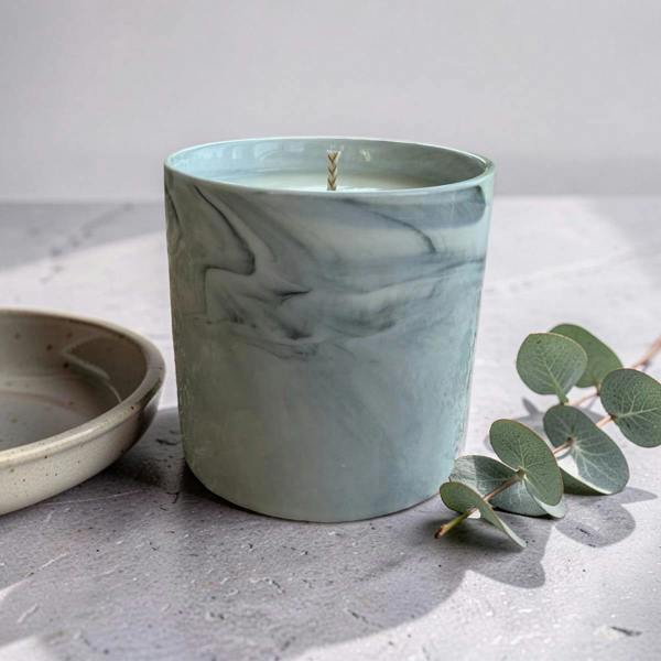 A marble-patterned teal candle with a single wick sits on a gray stone surface next to a ceramic dish and eucalyptus leaves.