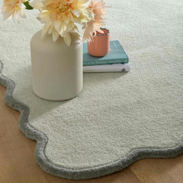 Light green rug with scalloped edge on a wooden floor, featuring a vase with flowers and books.
