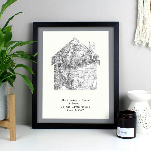 A black framed print showcasing an Ordnance Survey map cut into a house silhouette, personalised with a typed four-line quote, sitting on a white table.