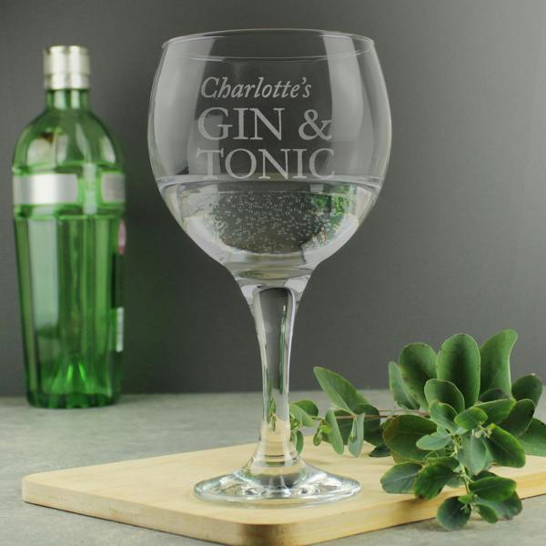 A personalised stemmed gin balloon glass by Abode Home & Gifts, featuring the custom script engraving "Charlotte's GIN & TONIC", filled with a clear drink and standing on a wooden serving board next to a green bottle and leafy eucalyptus sprigs.