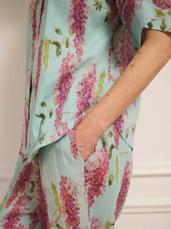 Woman wearing duck egg blue cotton gauze oversized pyjamas with pink wisteria floral print, short-sleeve button-through top and wide-leg trousers, standing barefoot indoors. Pocket detail